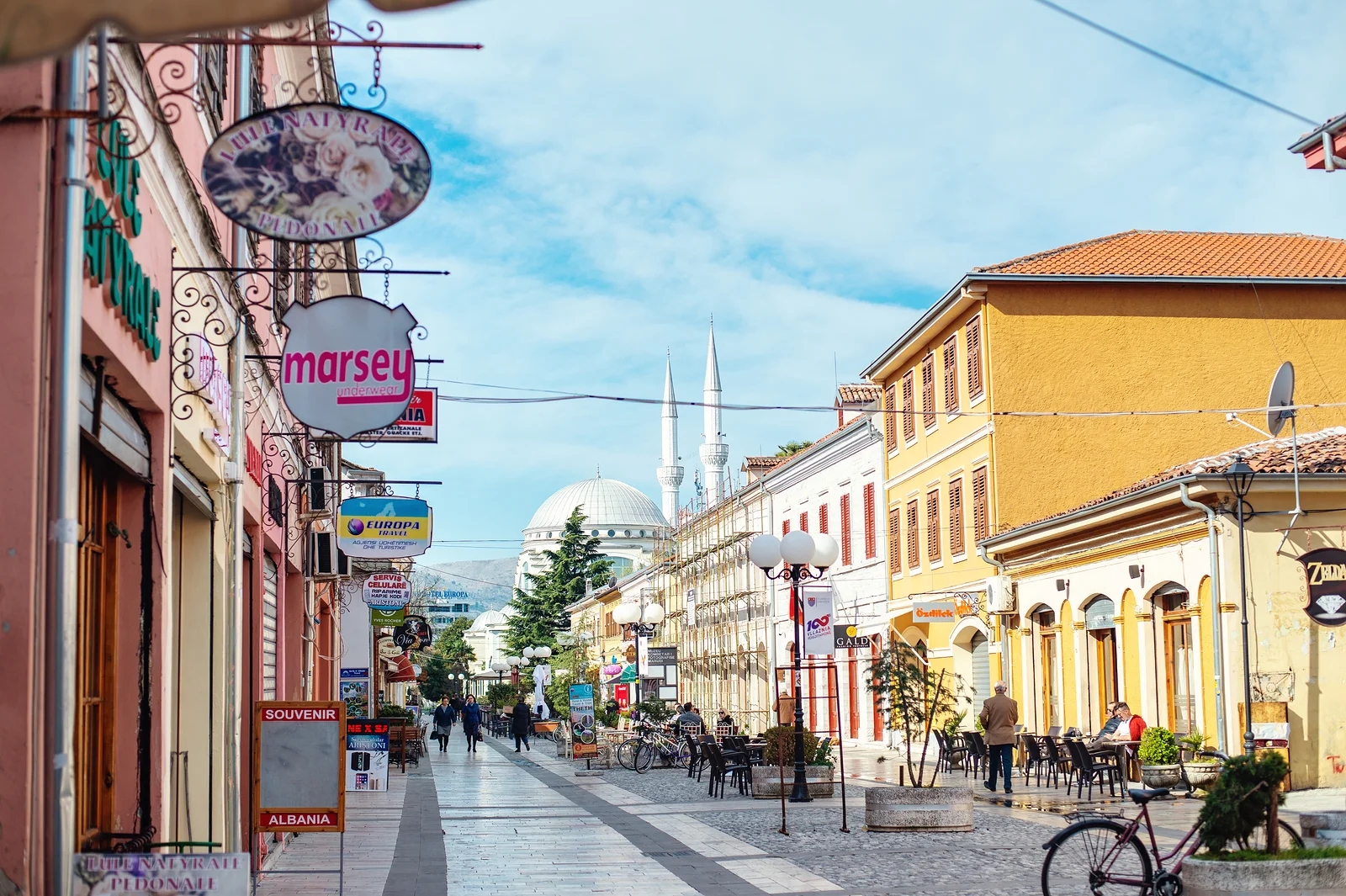 Pedestrian zone in central Shkodër with café terraces and the Ebu Bekr Mosque in the distance, two minutes on foot from NÓS Albania